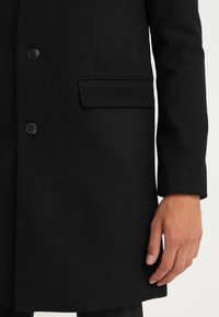 Cappotto di lana nera con taglio dritto, due bottoni e una tasca sul petto. La texture è liscia con una finitura opaca.