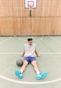 Scenă de teren de baschet cu o persoană așezată pe asfalt verde. Aceasta poartă un tricou fără mâneci de culoare gri și pantaloni scurți bleumarin, cu adidași turcoaz aproape.