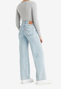 Person som bär ljusblå högmidjade Levi's jeans, grå räfflad långärmad topp och vita sneakers, står med ryggen vänd mot åskådaren.