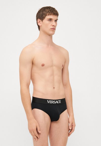 Svarte bomullsunderbukser med bred elastisk midjebånd som har hvit "VERSACE" merkevare. Glatt tekstur, tettsittende design, midjehøyde.
