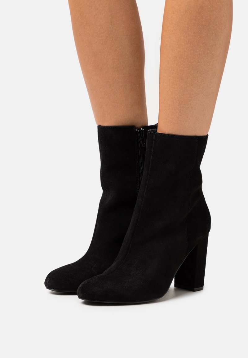 Femme portant des bottines en daim noir avec talons épais et fermetures éclair latérales, debout devant un fond clair uni.