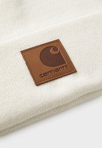 Strikket beige lue med en brun lærkvadratlapp som har et stilisert logo og merkevarenavnet "carhartt." Teksturert stoff.