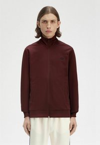 Giacca bordeaux con zip, colletto alto, tasche frontali e un logo discreto. Tessuto liscio e design aderente.