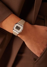 Montre numérique avec un cadran blanc hexagonal, un bracelet en silicone gris et des éléments en acier inoxydable. L'affichage indique l'heure et d'autres fonctionnalités.