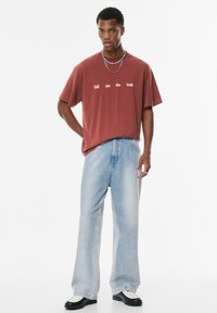 T-shirt graphique marron avec texte, jean en denim bleu clair ample et chaussures noires avec accents blancs, sur fond blanc uni.