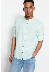 Camicia azzurra chiara con colletto alla coreana, maniche corte arrotolate e bottoni neri, indossata con jeans in denim blu.