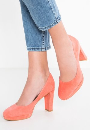 Füße, die korallenrote Wildleder-Pumps mit Blockabsatz tragen, kombiniert mit hochgekrempelten hellblauen Jeans auf weißem Hintergrund.