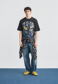 Νέος άνδρας στέκεται φορώντας μαύρο T-shirt με τύπωμα, δεμένο καρό πουκάμισο στη μέση, μπλε τζιν και κίτρινα αθλητικά παπούτσια σε μπλε χαλί.