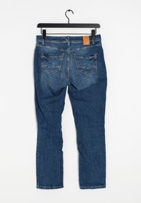 Siniset farkut mustassa henkariassa, takaa näkyvillä kaksi taskua ja ruskea nahkainen merkkilappu.