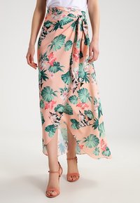 Žena v broskvově barevných květinových zavinovacích sukni s zelenými listy a růžovými květy, doplněná korálově zbarvenými sandály na kotníku a bílým topem.