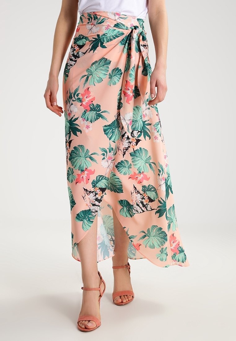 Žena v broskvově barevných květinových zavinovacích sukni s zelenými listy a růžovými květy, doplněná korálově zbarvenými sandály na kotníku a bílým topem.