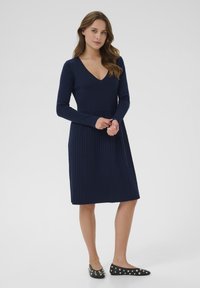 Femme aux longs cheveux bruns portant une robe longue à manches longues bleu marine à côtes et des ballerines noires cloutées, se tenant devant un fond clair uniforme.