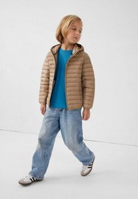 Beige pufferjack met horizontale stiksels, ritssluiting en capuchon; gedragen over een blauw shirt en gecombineerd met losse blauwe jeans en sneakers.