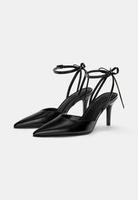 Décolleté nere in pelle con tacco alto, punta affilata, cinturino alla caviglia e dettaglio a fiocco. Texture liscia e design del tacco sottile.