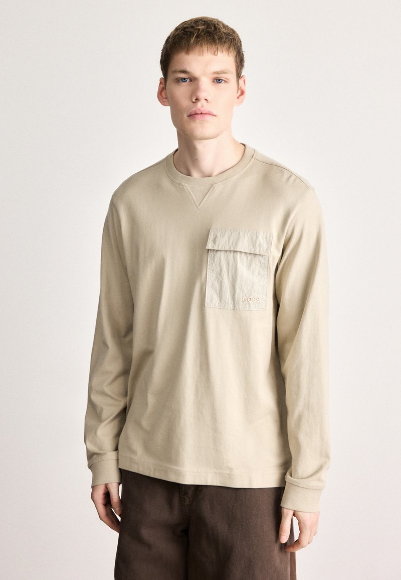 Béžová košeľa s dlhou rukávou, okrúhlym výstrihom a štruktúrovanou náprsnou vreckovou na ľavej strane; mäkká bavlnená látka; voľný strih.