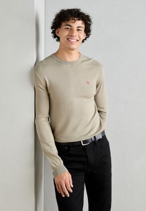 Beige long-sleeve sweater with ribbed cuffs, featuring a small red logo. Paired with black jeans and a black belt. Smooth texture.