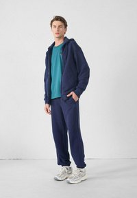 Sweatshirt à capuche bleu marine avec une fermeture éclair à l'avant et des poches, associé à un pantalon fuselé. T-shirt turquoise en dessous. Chaussures de sport grises.