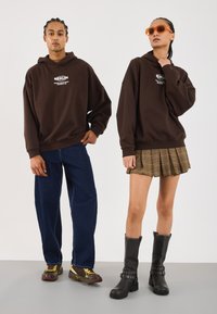 Bruine oversized hoodie met witte grafische print; gecombineerd met donkerblauwe wijde jeans. Vrouw in een geruite rok en zwarte kniehoge laarzen. Oranje zonnebril.