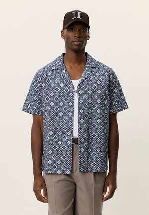 Kortærmet skjorte med knapper i blå og hvidt blomsterprint, med camp-krave og løs pasform, kombineret med beige bukser og en kasket.