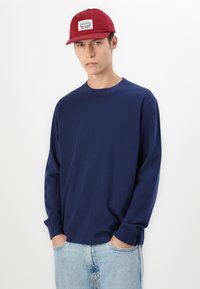 Granatowy sweatshirt z długimi rękawami i okrągłym dekoltem, w połączeniu z czerwoną czapką z naszywką logo. Lekko niebieskie dżinsy dopełniają całość.