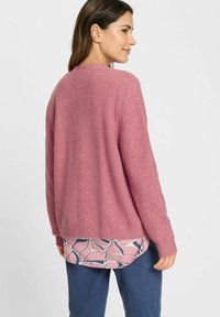 Pink fuzzy sweater with a relaxed fit and ribbed cuffs, layered over a patterned pink and blue blouse, paired with blue pants.