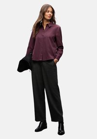 Burgunderfarbene Bluse mit Kragen, kombiniert mit lockeren, dunkelgrauen Hosen und schwarzen Stiefeletten. Das Modell hält eine schwarze Tasche.