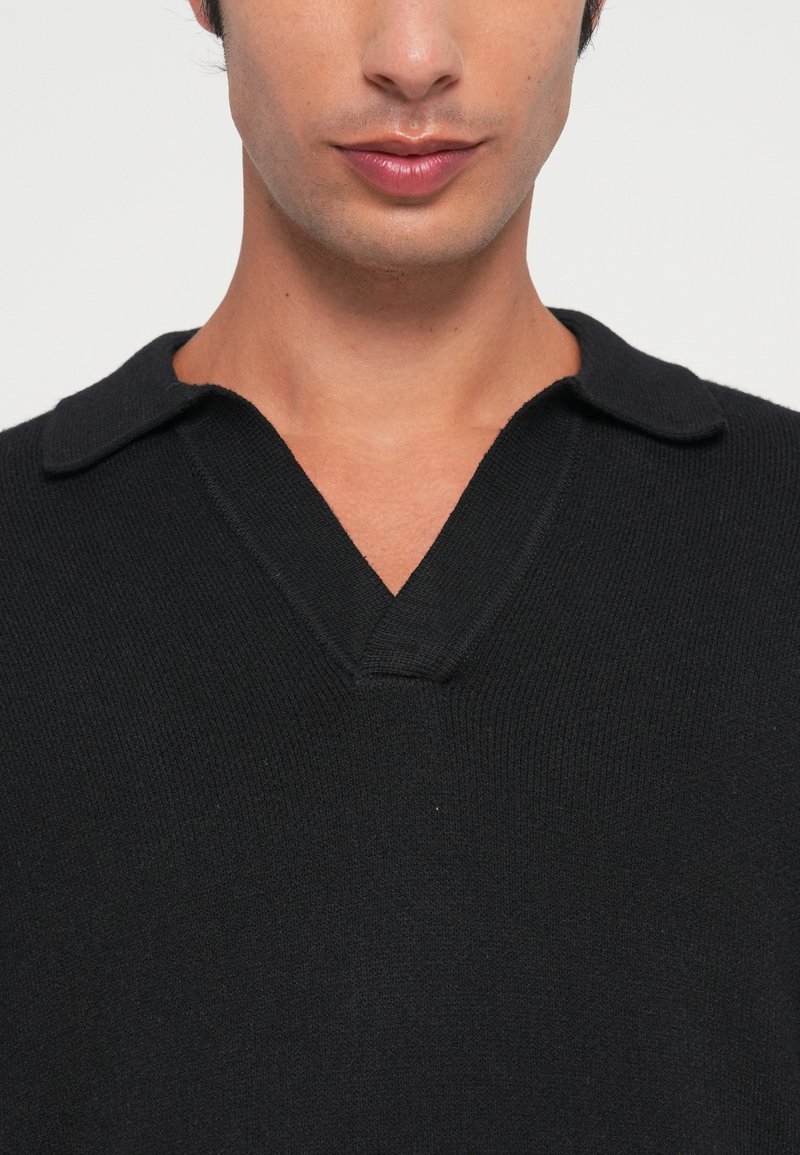 Giovane uomo che indossa un maglione a maglia nero con scollo a V e colletto ripiegato, mostrato dalla zona del collo al petto.