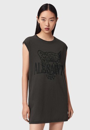 Junge Frau mit langen dunklen Haaren, die ein ärmelloses schwarzes Kleid trägt, das einen brüllenden Leoparden und den Text "ALESSANDR" zeigt.