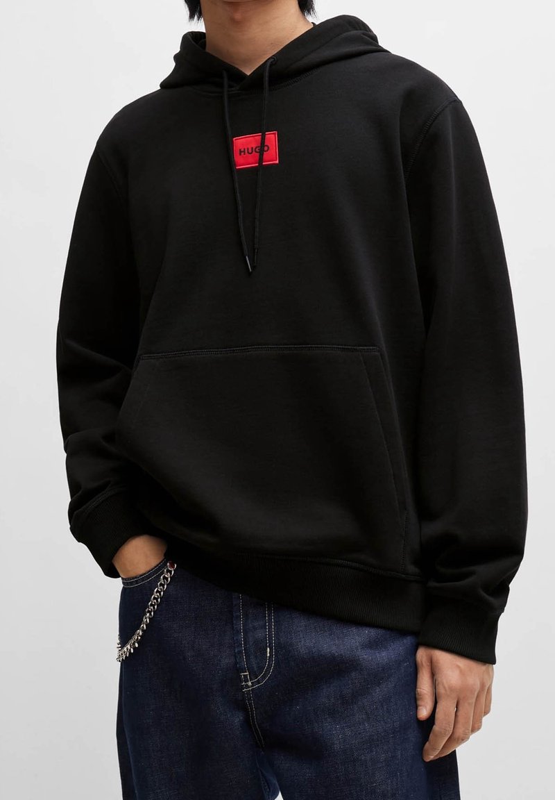 Personne portant un sweat à capuche noir avec une étiquette rouge « HUGO » sur la poitrine et un jean bleu foncé avec une chaîne argentée attachée à la poche.