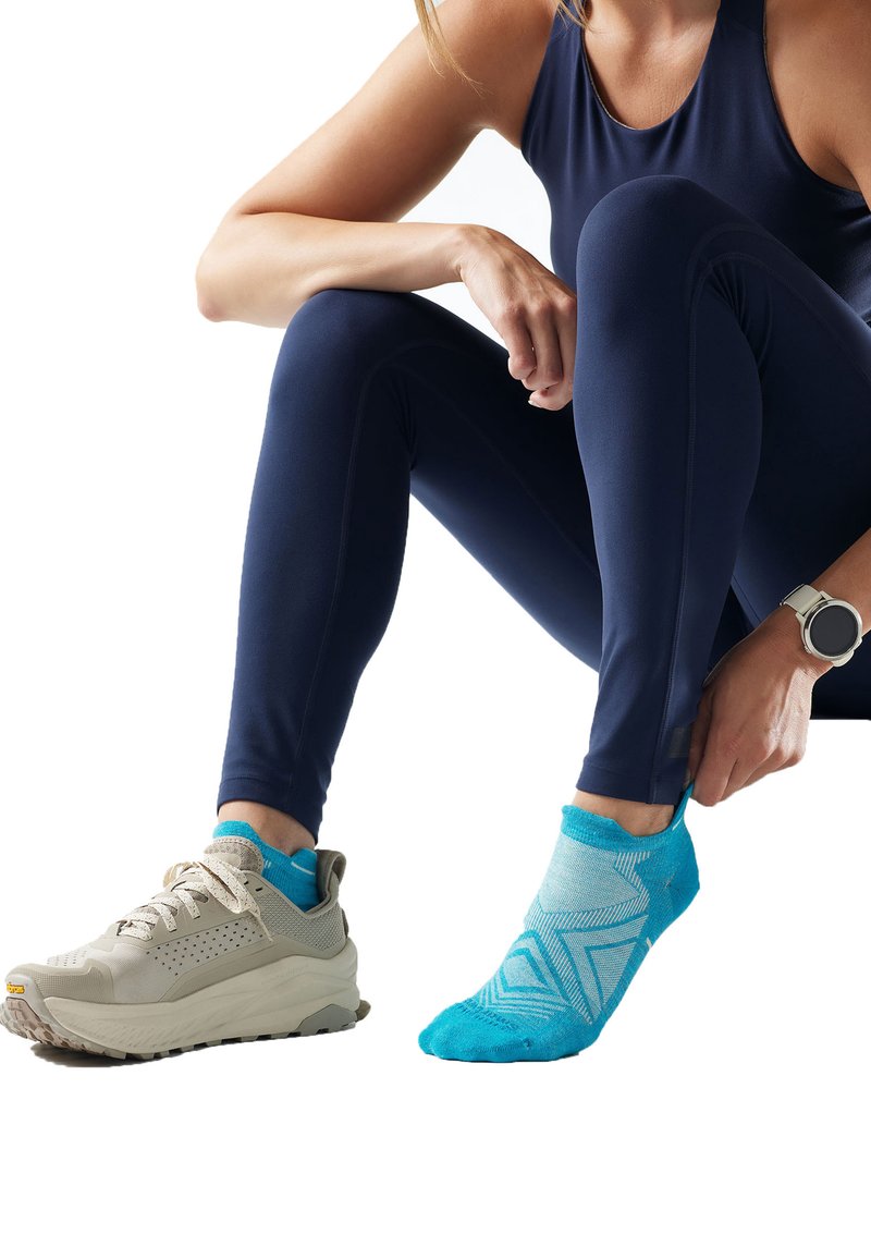 Personne portant des vêtements de sport bleu marine et une montre connectée remonte une chaussette bleue à motifs en étant assise, avec une chaussure beige à l'autre pied.