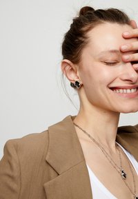 Pendientes negros y plateados con diseño floral, usados por una persona con una blazer beige, collar plateado y camiseta blanca. Piel con ligeras pecas.