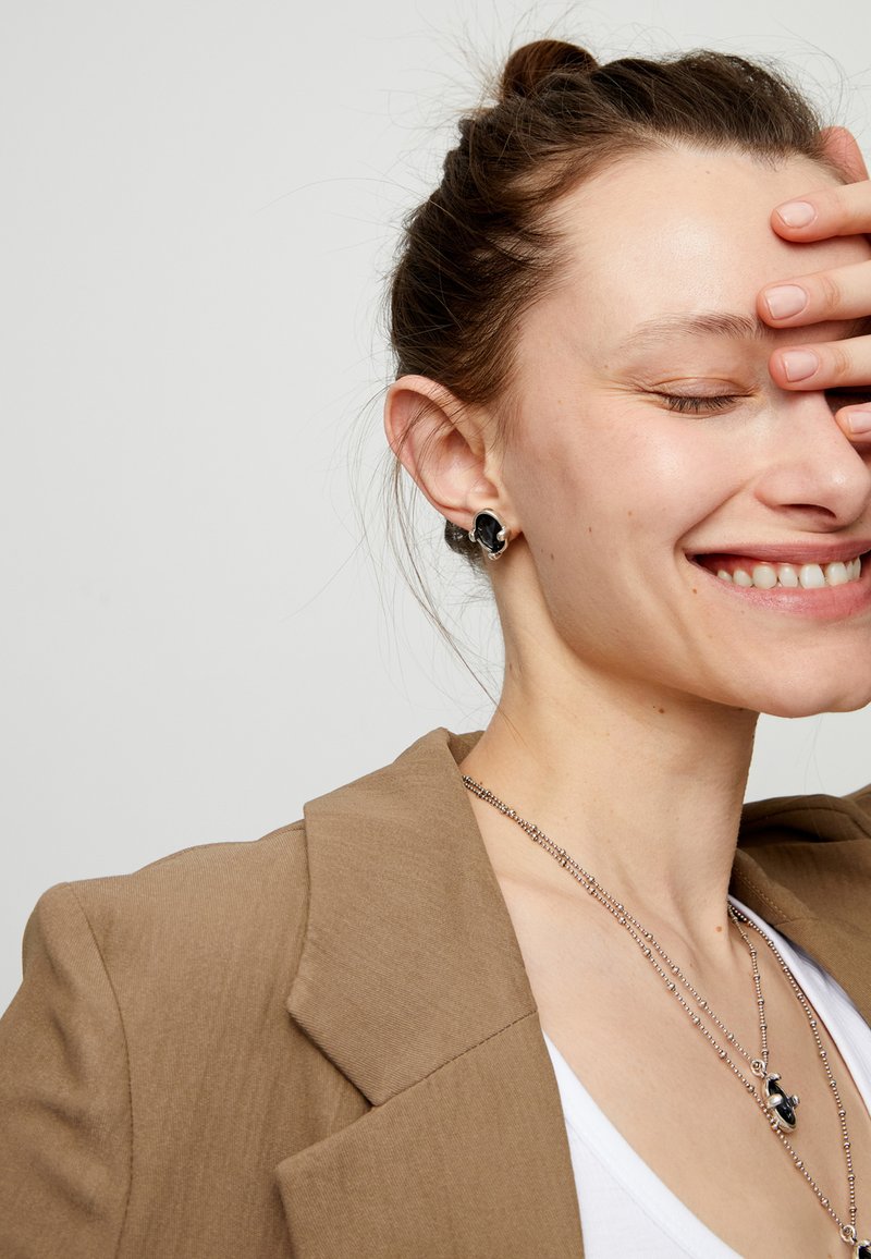Pendientes negros y plateados con diseño floral, usados por una persona con una blazer beige, collar plateado y camiseta blanca. Piel con ligeras pecas.