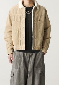 Beige kordjacka med en vit pälskrage, med framfickor och tryckknappar, bärs över en svart strukturerad tröja och grå cargobyxor.