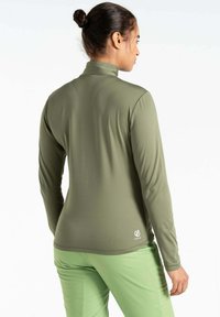 Langärmliges Oberteil in olivgrün, glatte Textur, hoher Kragen, figurbetontes Design. Logodetail unten rechts. Kombiniert mit hellgrünen Hosen.