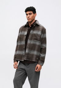 Καφέ φανελένιο σακάκι πουκαμίσου με γκρι καρό σχέδιο, κουμπιά με πίεση και κομμένο γιακά. Φορεμένο πάνω από καστανό πουκάμισο και συνδυασμένο με γκρι παντελόνι.