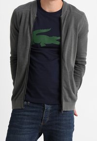 Cardigan grigio con zip sopra una t-shirt blu navy con un grafico di coccodrillo verde. Indossato con jeans blu. Tessuto liscio e design aderente.
