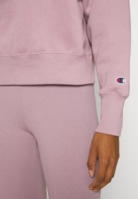 Felpa viola chiaro con polsini e orlo a coste, abbinata a leggings viola chiaro coordinati. Presenta una patch con logo sulla manica della felpa.