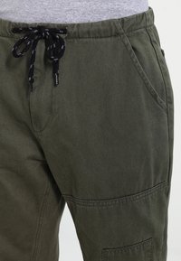 Pantalones cargo verdes hechos de tela duradera, con cintura de cordón en negro, bolsillos laterales y acabado texturizado.