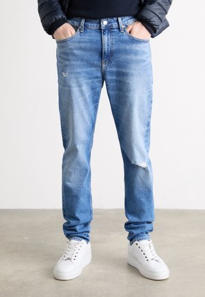 Mann trägt hellblaue, abgenutzte Jeans, weiße Sneakers, schwarze Jacke, Hände in den Taschen, steht auf einem grauen Boden mit schlichtem weißen Hintergrund.