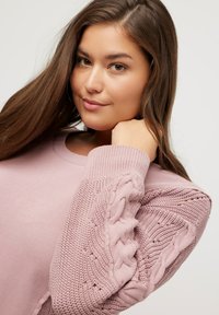 Vaaleanpunainen neuletakki, jossa on sileä vartalo ja raketinkuvioidut hihat, joissa on yksityiskohtaisia kuvioita. Pyöreä pääntie ja joustavat hihansuut.