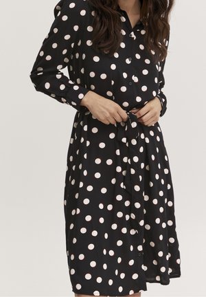 Kobieta poprawiająca pasek w talii czarnej sukienki na guziki z dużymi białymi grochami i długimi rękawami.