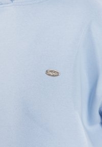 Sudadera azul claro con textura suave y un pequeño parche de logo plateado en la parte delantera, con un diseño simple y casual.