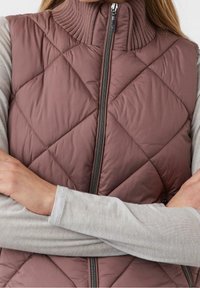 Femme portant un gilet matelassé mauve avec un col côtelé haut sur une chemise à manches longues gris clair, les bras croisés sur la poitrine.