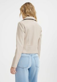 Giacca cropped in pelle beige con colletto classico e design minimalista. Presenta spalle e schiena senza cuciture, abbinata a jeans blu chiaro.