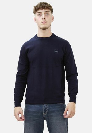 Maglione a maniche lunghe blu navy in un tessuto a maglia morbido, con collo rotondo e piccolo logo sul petto. Indossato con jeans blu.