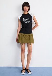 Czarny t-shirt graficzny z białymi rękawami, w połączeniu z khaki spódnicą z metalowymi elementami i widocznymi postrzępionymi brzegami, noszoną z czarnymi butami.