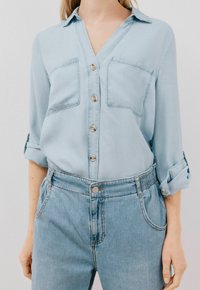 Camicia di denim azzurro chiaro con scollatura a V, due tasche sul petto e chiusura con bottoni. Abbinata a jeans azzurro chiaro a vita alta con cintura elastica.