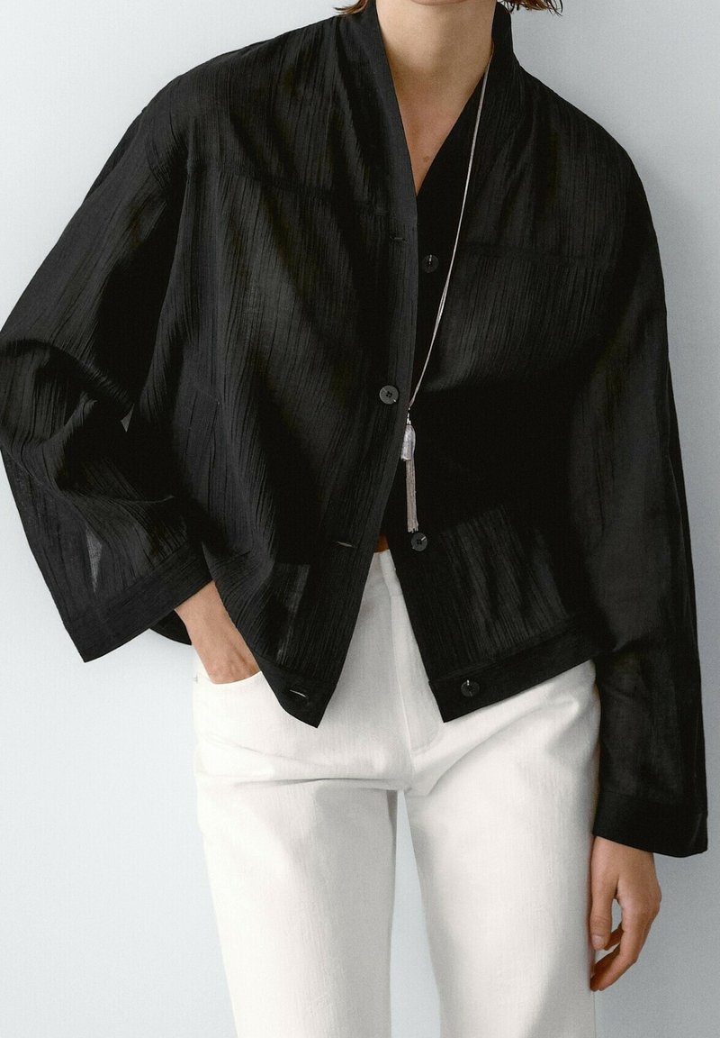 Black cropped jacket with a textured finish, featuring a shawl collar, front buttons, and wide sleeves, paired with white pants.