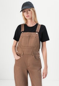 Bruine denim tuinbroek met een gestructureerde pasvorm, grote frontzak en verstelbare banden. Gekocht met een zwart T-shirt en een donkere pet.