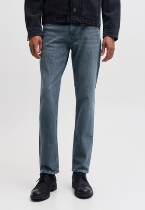 Homme portant un jean bleu, une veste noire et des chaussures noires à lacets, debout devant un fond uni.